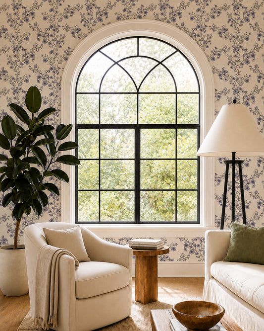 Blossom Whisper Dusty Blue floral wallpaper in a living room with an arched window, cream seating and natural wood accents.